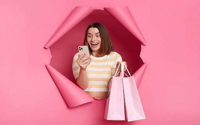 A woman comparing prices on her phone while holding a Victoria's Secret shopping bag, suggesting price matching.